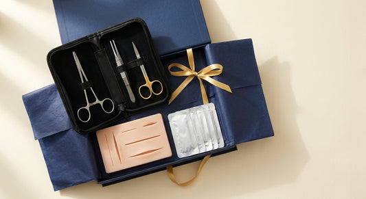 Navy gift box with gold ribbon, open to reveal a surgical instrument case, silicone suture pad and suture packets