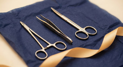 Close-up of stainless steel needle holder, forceps and iris scissors on navy tissue with gold ribbon